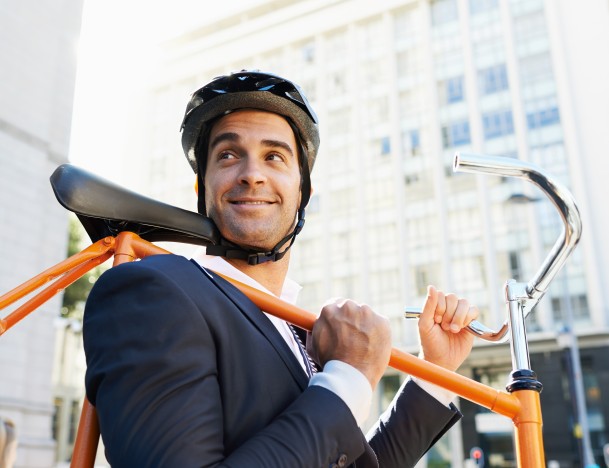 Mann im Anzug mit Fahrradhelm trägt sein Fahrrad mit der Stange über der Schulter – Symbolbild für eine Unfall invest Versicherung